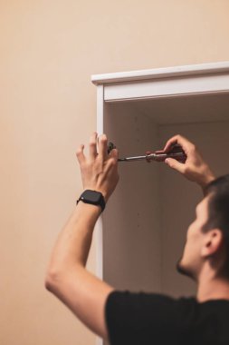 Genç, beyaz tenli, esmer adam, yatak odasındaki dolapta dikilirken metal bir tornavidayla düzüşüyor. Mobilya montaj konsepti.