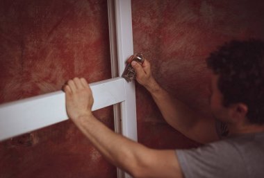 Kıvırcık saçlı genç beyaz bir adam pencere kenarındaki çatlakları inşaat bıçağıyla temizliyor ve onları montaj için hazırlıyor. Ev yenileme, montaj konsepti