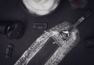 Ortasında traş köpüğü lekesi olan siyah bir arka planda jilet, tarak, bir şişe tıraş losyonu, bir kase köpük ve fotokopi alanı olan elektrikli tıraş bıçağı, yakın plan. Konsept: