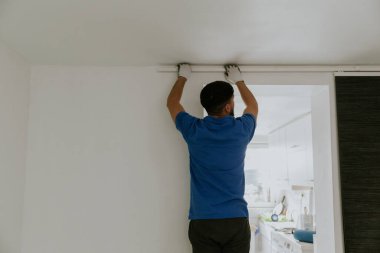 Mavi tişörtlü, beyaz tenli genç bir adam arkadan dikiliyor ve kapıdaki duvardan koyu gri, modern kağıt perdeleri kaldırıyor..