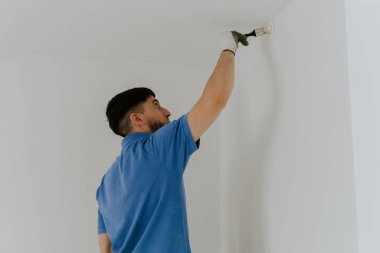 Mavi tişörtlü esmer, yakışıklı beyaz bir adam yarım yamalak duruyor ve duvarların köşelerini küçük bir fırça ile beyaz boyayla boyuyor..