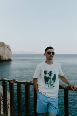 Güneşli bir yaz akşamı, güneşli bir yaz akşamı, güneşli elbiseli yakışıklı bir esmer, ahşap bir terasta denizin zeminine ve kayalara yaslanır..