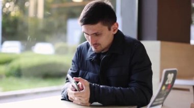 Koyu renk ceketli, beyaz tenli genç bir adam bir lokantanın penceresinin yanındaki masada oturur ve dikkatlice telefon ekranına bakar. Parmaklarını ekrandan geçirir.