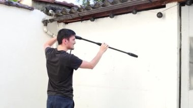 Arkadan gelen beyaz tenli genç bir adam bir yaz akşamı evinin arka bahçesinde bir Karcher 'la birlikte bir bahçe evinin beyaz duvarlarını yıkıyor..