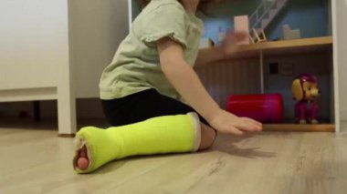 Sol bacağında açık yeşil alçı olan küçük, güzel bir beyaz kız oyuncaklarla dolu bir odada yerde otururken büküldü..