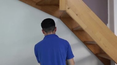 Mavi tişörtlü, siyah saçlı beyaz bir adam arkada duruyor ve salonun ahşap merdivenlerinin altına küçük bir fırça ile beyaz bir duvar boyuyor..