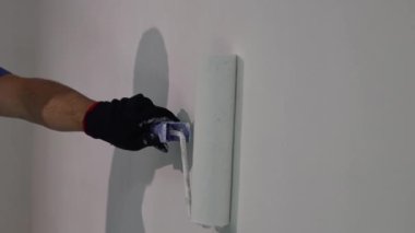Tanımlanamayan siyah eldivenli genç bir beyaz adam bir odada yuvarlak silindirli, yakın çekimde beyaz boyalı bir duvar boyuyor..