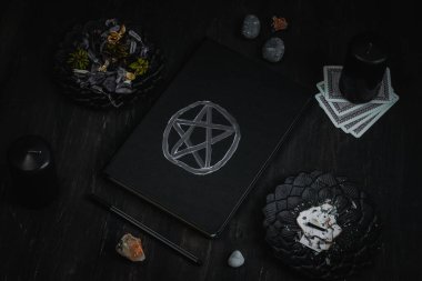 Sihirli beyaz sembollü siyah bir deri kitap, siyah mumlar, siyah metal tabaklar, kurutulmuş çiçekler, ritüel sopaları, tarot kartları, siyah ahşap arka planda bir kalem ve taşlar, yakın plan üst manzara. Esrarengiz kavram, karanlık tarz..