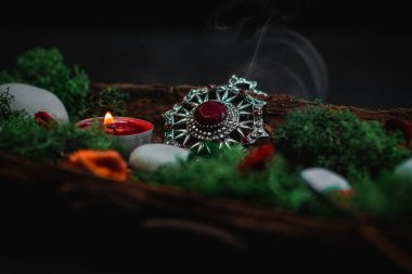 A ritual amulet with a red gem, green moss, a candle, a dried flower, streaming smoke and gray stones in the bark of a tree lies on a black background, close-up side view. Esoteric concept, dark style.