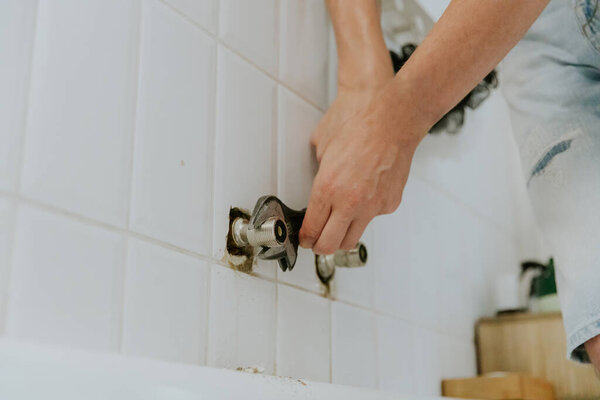One young Caucasian unrecognizable man manually tightens the nut from the faucet with two hands with an adjustable wrench in the bathroom sticking out of the wall, standing in the bathroom during the