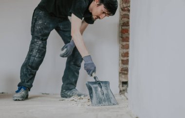Siyah üniformalı, yakışıklı, beyaz bir kadın ayakta duruyor, eğiliyor, yerden kürekle yıkıntılar topluyor, yakından bakıyor..