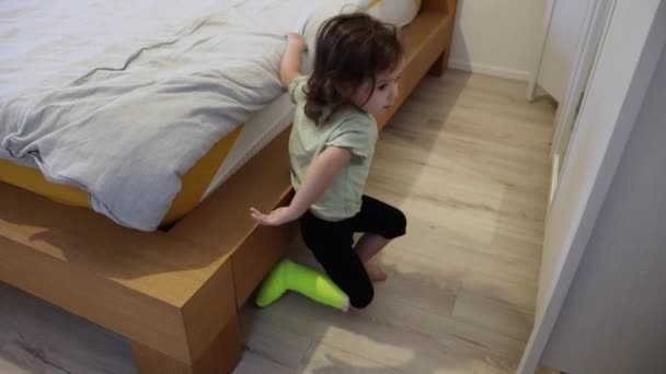 一个白种人的小女孩 左腿上戴着浅绿色的石膏 从卧室的大床上跳到地板上 跪了下来 开始玩玩具 近距离侧视 — 图库视频影像