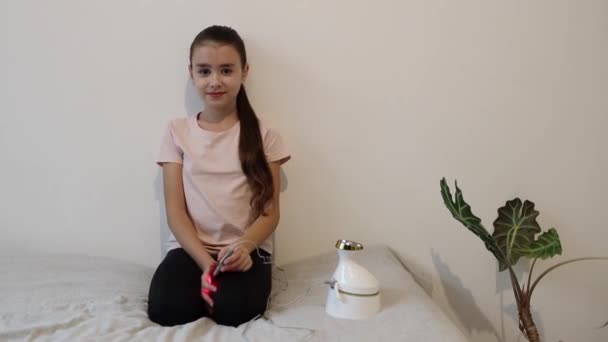 一位漂亮的 快乐的白种人小女孩把一根有红外光的棍子放在她的右耳里进行治疗 她正在为感冒热身 坐在靠近儿童医院白墙的床上 — 图库视频影像
