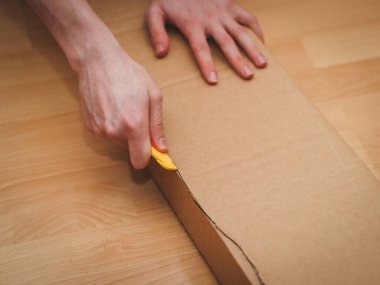 Kafkas bir gencin elleri, odasının zemininde otururken, uzun, dikdörtgen bir karton kutuyu sarı bir inşaat bıçağıyla kesiyor. Paket teslimatı kavramı