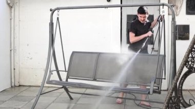 Koyu renk elbiseli esmer, beyaz bir adam, bir yaz akşamı evinin küçük arka bahçesinde güçlü bir su basıncıyla bir salıncağın koltuklarını yıkıyor.