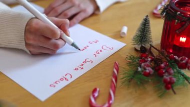 Beyaz kazaklı, tanınmayan beyaz bir genç kız, Noel Baba 'ya Noel süslemeleri olan ahşap bir masada oturan kırmızı keçeli kalemle bir mektup yazar.