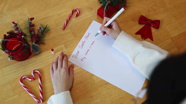 白いセーターで認識できない白いコーカサスの十代の少女は クリスマスの装飾と木製のテーブルに座っている間 赤いフェルトチップペンで親愛なるサンタクロースに希望の手紙を書いています ダイニング — ストック動画