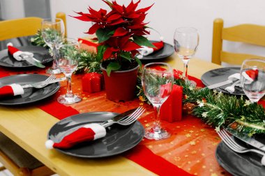 Bir Noel çiçeği tencerede duruyor gri tabaklar, bıçaklar, çatallar, bardaklar, mini Noel Baba şapkaları, kırmızı masa örtüsü ve mutfaktaki bir kış akşamında yeşil süslemeler