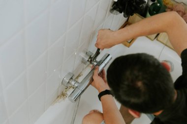 Kafkasyalı genç bir esmer adam, küvetin kenarında otururken ayarlanabilir bir anahtar kullanarak musluğun içindeki cevizi elle sıkıyor. Adım adım..