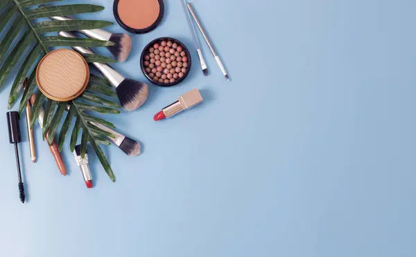 A set of cosmetics from a face powder box, red lipsticks, makeup brushes, mascara and pencils for lips and eyebrows with a palm branch on the left on a blue background with copy space on the right