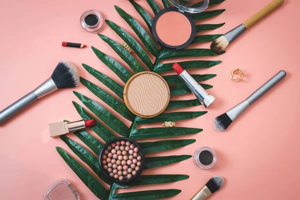 A set of cosmetics from face powder boxes, makeup brushes, red lipstick, eye shadow and rings on a palm tree branch lie diagonally on a pink background, flat lay close-up of sharpness. The concept of
