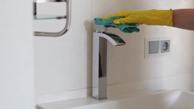 Sarı eldivenli beyaz bir kadının elleri banyoda yıkamayı bitirdikten sonra uzun bir lavabo musluğundan suyu yeşil bir mikrofiber bez ile siliyor.