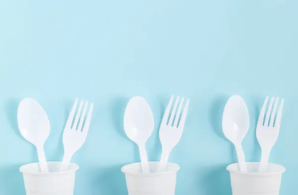 Plastic white glasses with spoons and forks inside them lie in a row from below on a light blue background with copy space from above, flat lay close-up. Ecology concept, disposable cutlery.