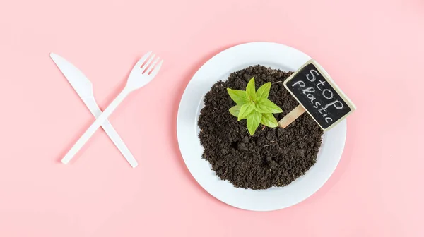 Plastic knife and fork crossed, white plate with black earth, green sprout and peg with the inscription: stop plastic lying on a light pink background, flat lay close-up. The concept of ecology and