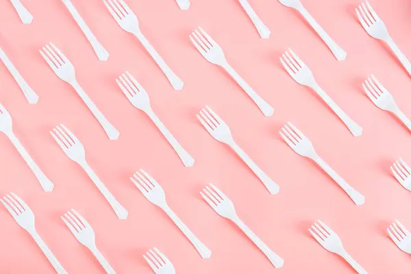 A lot of white plastic forks lie diagonally on a light pink background, flat lay close-up. The concept of ecology, plastic garbage and disposable plastic tableware.