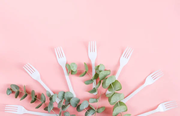 White plastic forks with green branches and leaves fan out from below on a pink background with little copy space on top, flat lay close-up. The concept of ecology and plastic waste, disposable