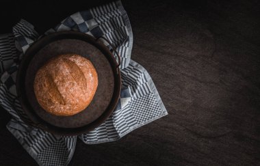 One round rye bread in a vintage metal plate with a kitchen napkin lies on the left on a black stone background with copy space on the right, flat lay close-up. Bread baking concept.