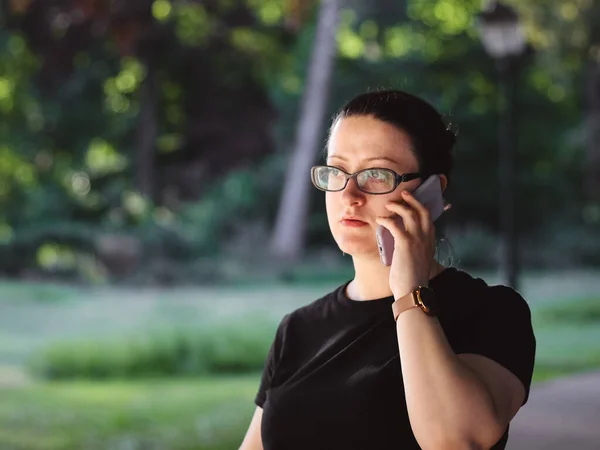 Caucasian woman in glasses and a black t-shirt emotionally talking on a mobile phone waving her hand in a public park against the background of blurry trees, close-up side view. Concept of using a
