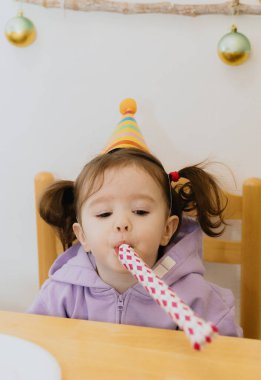 Konik şapkalı güzel bir beyaz kız ve kafasında iki at kuyruğu olan bir şenlik masasında oturur ve güçlü bir şekilde kağıt boruya üfleyerek yan tarafa bakar, doğumgününü kutlar, yakın çekimde yandan bakar.