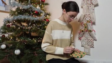 Güzel bir beyaz kız, duvar takviminin cebinden hediyesini açar ve onu zevkle inceler. Zarif bir Noel ağacının arka planında oturur..
