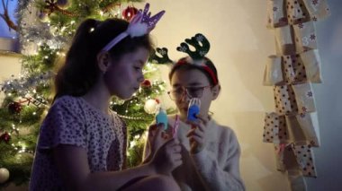 İki güzel beyaz kız mutlu bir şekilde şeker yer ve duvar takvimindeki oyuncaklarla oynar, akşamları zarif bir Noel ağacının arka planında otururlar, yakın çekimde..