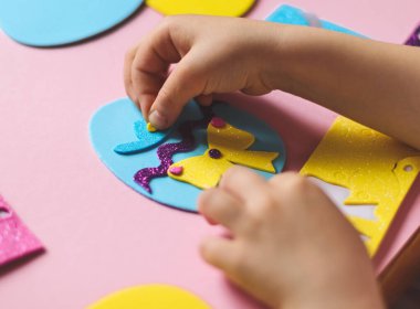 Küçük beyaz bir kızın elleri, parmaklarıyla mavi bir yumurtanın üzerinde sarı bir keçeli kalem etiketi, şapkası ve leylak zigzag ile çocuk masasında oturuyor.