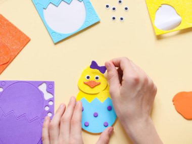 Beyaz bir genç kızın elleri sarı bir tavuğun üzerine leylak bir fiyonk yapıştırıyor parmaklarıyla hissettiği, bir çocuk masasında oturuyor soluk sarı bir arka planda el yapımı eşyalar ve