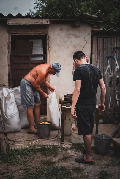 Two Caucasian men, old and young, are manually making dry compound feed for pets in a homemade crusher, standing on the street of a village house in the countryside, close-up side view in dark style