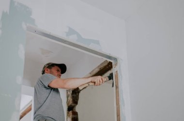 Gri tişörtlü ve şapkalı genç beyaz bir adam, bir eliyle balta kullanarak, aşağıdan seçici bir odak ile eski macunu kapı eşiğinden temizliyor. İnşaat işi kavramı.