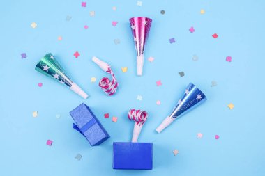 Renkli borular, ıslıklar ve dağınık kağıt konfetilerin döküldüğü açık mavi bir hediye kutusu mavi arka planda, düz bir yakın plan yatar.