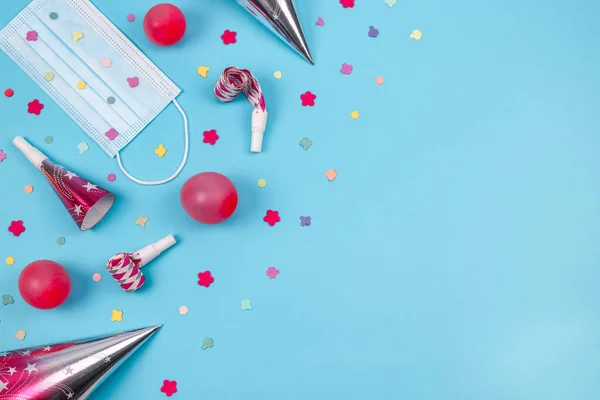 Inflated pink three balloons, medical mask, scattered confetti paper, colorful whistles and birthday cone hat lie on the left on a blue background with copy space on the right, flat lay close-up.