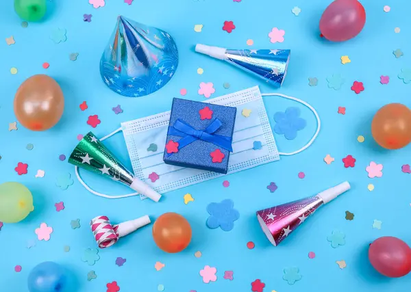 One cone birthday hat, whistle, shiny gift box, scattered paper confetti, inflated multi-colored balls and a medical mask with felt decorative flowers around lie on a blue background, flat lay close-up.