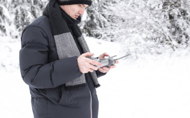 A young handsome caucasian man in a blue down jacket and a gray knitted hat controls a drone through a gray remote control in his hands while standing on a snowy meadow in a winter forest reserve in Belgium, close-up side view. The concept of winter 