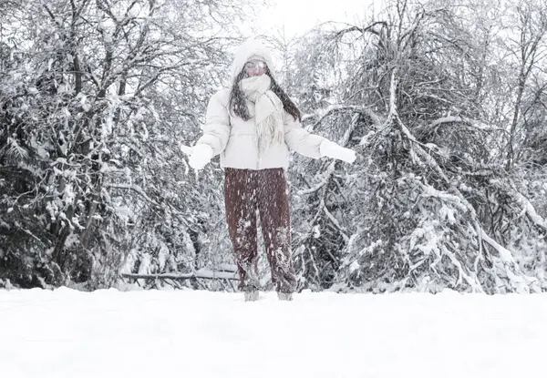A young girl in a white down jacket with a hood and brown pants throws up snow covering her eyes, standing in a winter forest in a nature reserve in Belgium, close-up side view.Using technology concept, winter holidays.