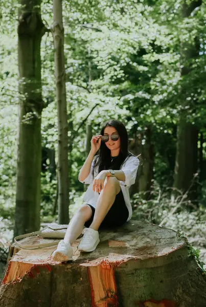 stock image One beautiful young Caucasian brunette girl in sunglasses and stylish clothes poses while sitting on a large sawn stump in the park on a sunny spring day, close-up side view.