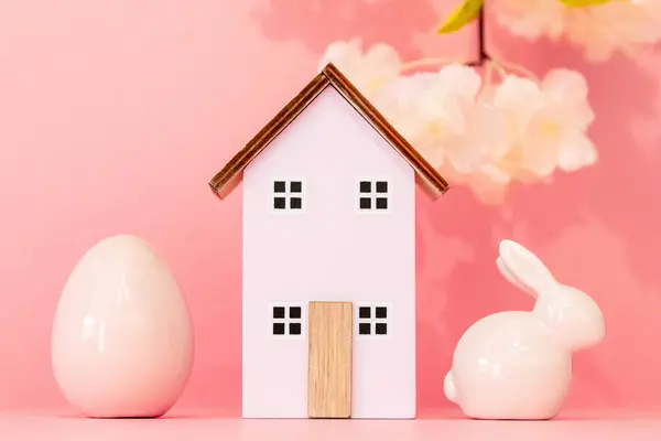 One wooden house with a porcelain Easter egg and a bunny stand on a pink background with a blurred branch of apple tree flowers, close-up view from below with selective focus.
