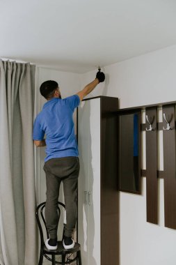 Mavi tişörtlü esmer, beyaz tenli bir genç tavanın kenarlarını beyaz boyayla boyuyor. Küçük bir fırça kullanıyor. Sırtı pencerenin yanındaki sandalyede duruyor.