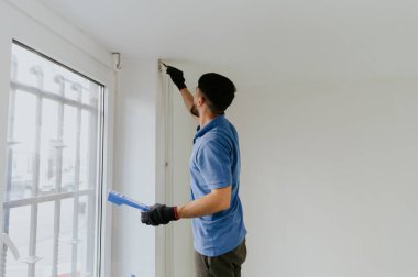 Koyu renk sakallı, mavi tişörtlü, genç bir beyaz adam pencerenin kenarında duruyor ve duvarın köşelerini beyaz boyalı, yakın plan bir fırçayla boyuyor.