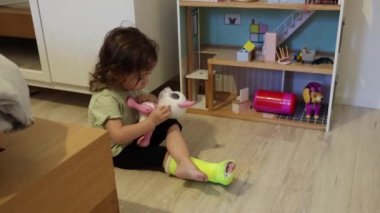 Sol bacağında açık yeşil alçı olan küçük, güzel bir beyaz kız odada yerde oturuyor ve parlayan gözleri de dahil olmak üzere çocuk evindeki plastik kedi oyuncağıyla oynuyor.