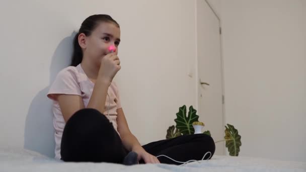 一个漂亮的白种人小女孩拿着一根带有红外光的手杖 在左鼻道上做菜 靠着墙坐在床上 两腿交叉 — 图库视频影像